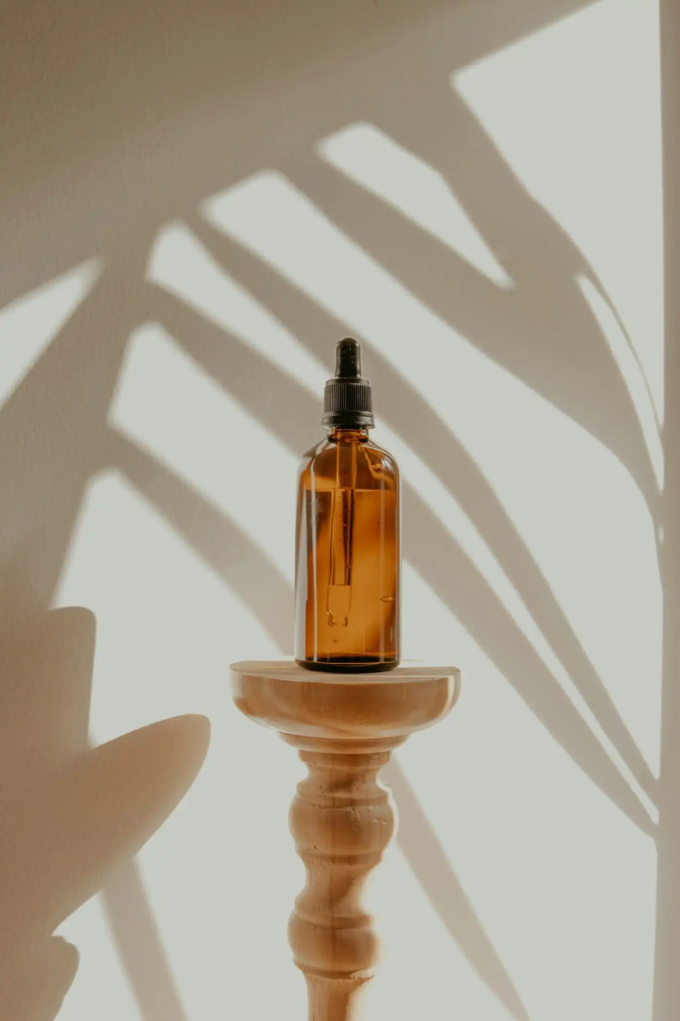 An amber glass dropper bottle on a turned wooden stand, with the shadow of palm leaves on the wall