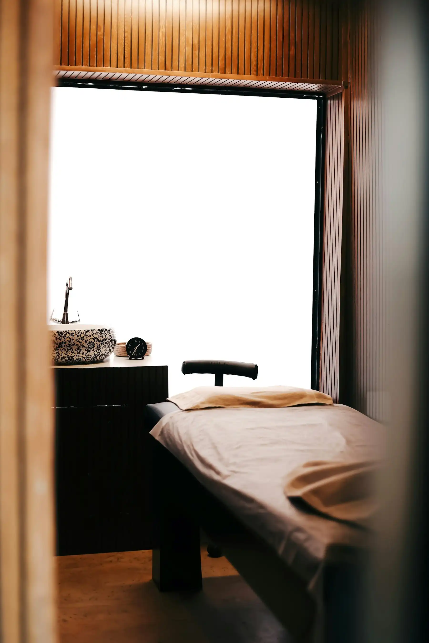 A quiet treatment room with a made-up treatment bed, warm wood paneling and soft daylight from a tall window